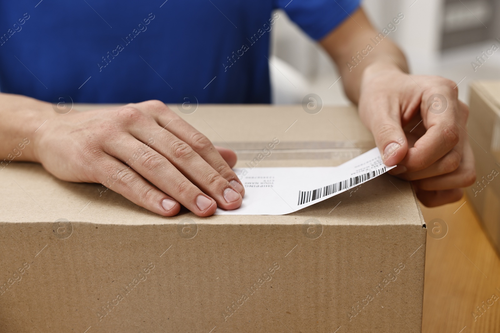 Photo of Man sticking shipping label with barcode on parcel at table indoors, closeup