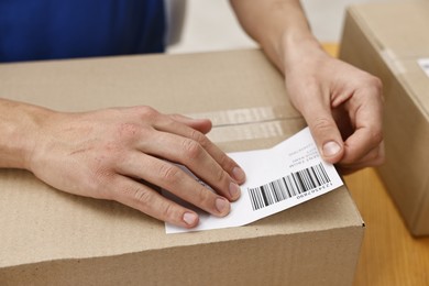 Photo of Man sticking shipping label with barcode on parcel at table indoors, closeup