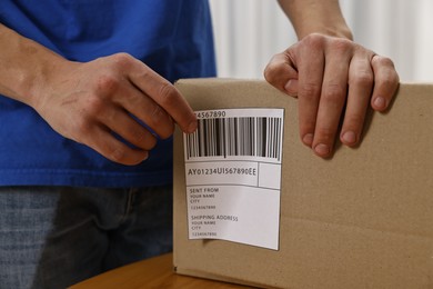 Photo of Man sticking shipping label with barcode on parcel at table indoors, closeup