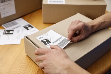 Photo of Man sticking shipping label with barcode on parcel at wooden table indoors, closeup