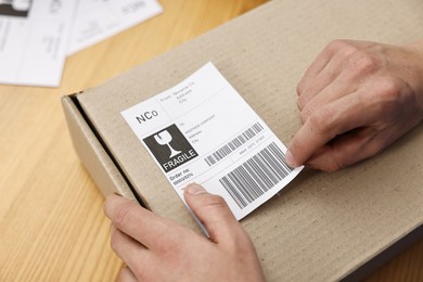 Photo of Man sticking shipping label with barcode on parcel at wooden table indoors, closeup