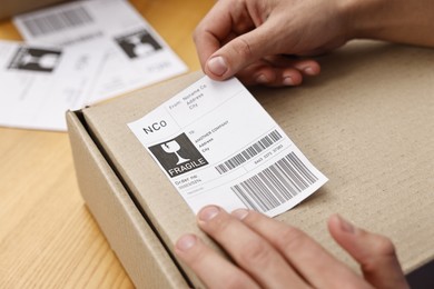 Photo of Man sticking shipping label with barcode on parcel at wooden table indoors, closeup