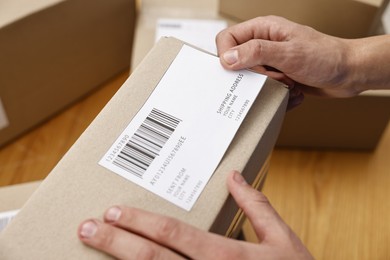 Photo of Man sticking shipping label with barcode on parcel at table indoors, closeup