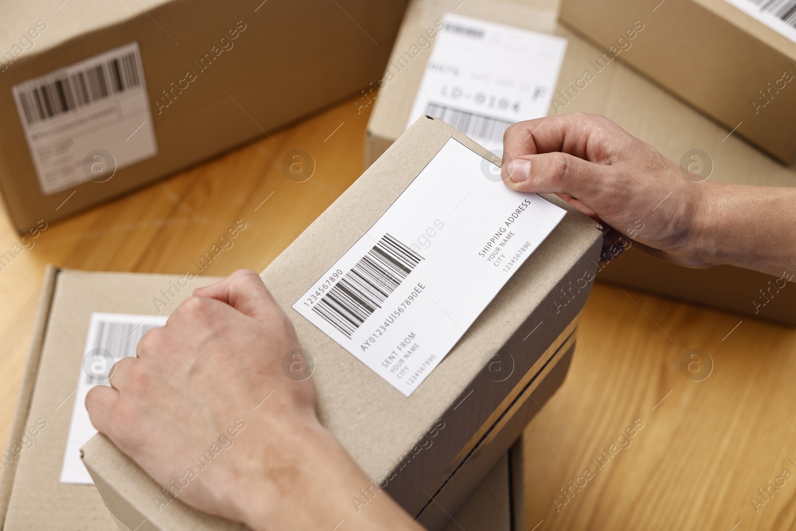 Photo of Man sticking shipping label with barcode on parcel at wooden table indoors, above view