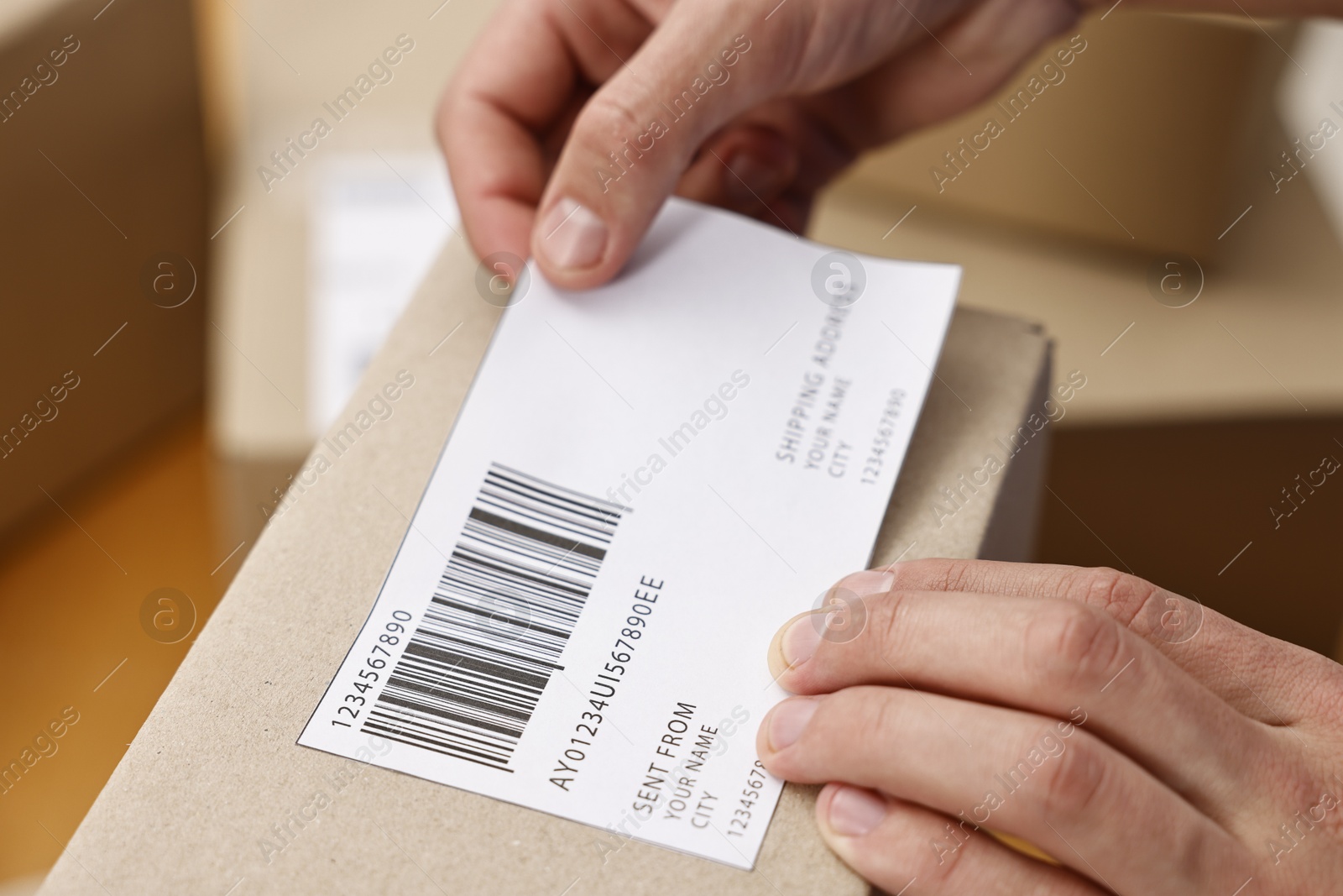 Photo of Man sticking shipping label with barcode on parcel indoors, closeup