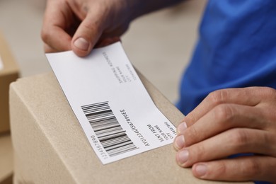 Photo of Man sticking shipping label with barcode on parcel indoors, closeup