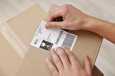 Man sticking shipping label with barcode on parcel indoors, closeup