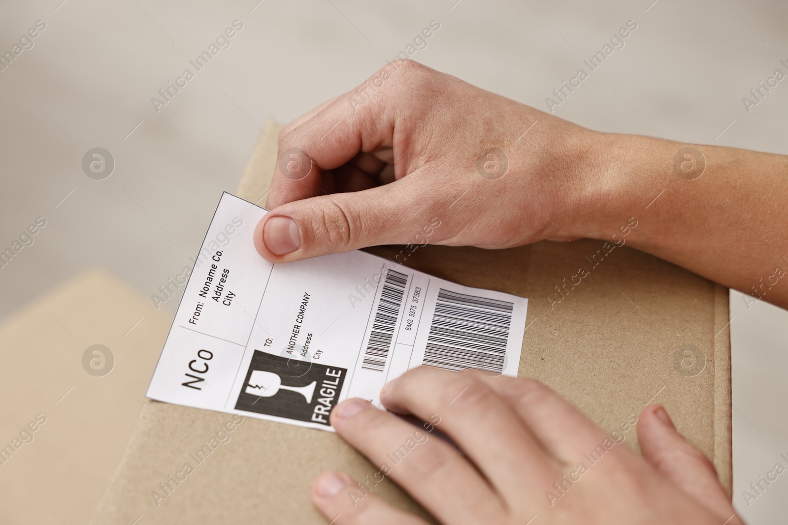 Photo of Man sticking shipping label with barcode on parcel indoors, closeup