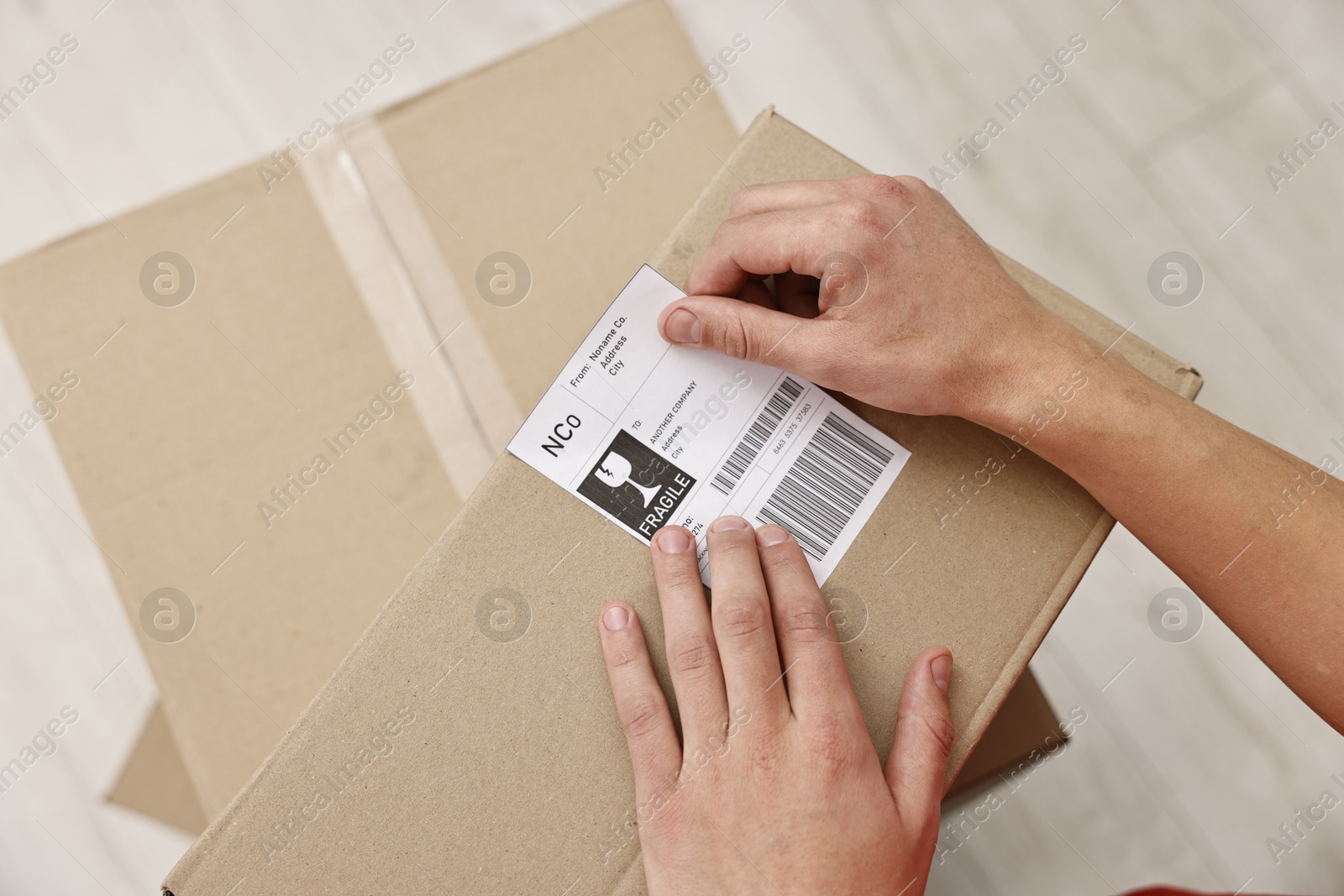 Photo of Man sticking shipping label with barcode on parcel indoors, above view