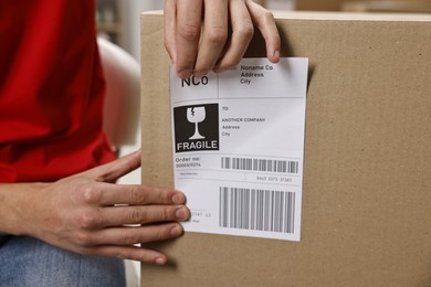 Photo of Man sticking shipping label with barcode on parcel indoors, closeup