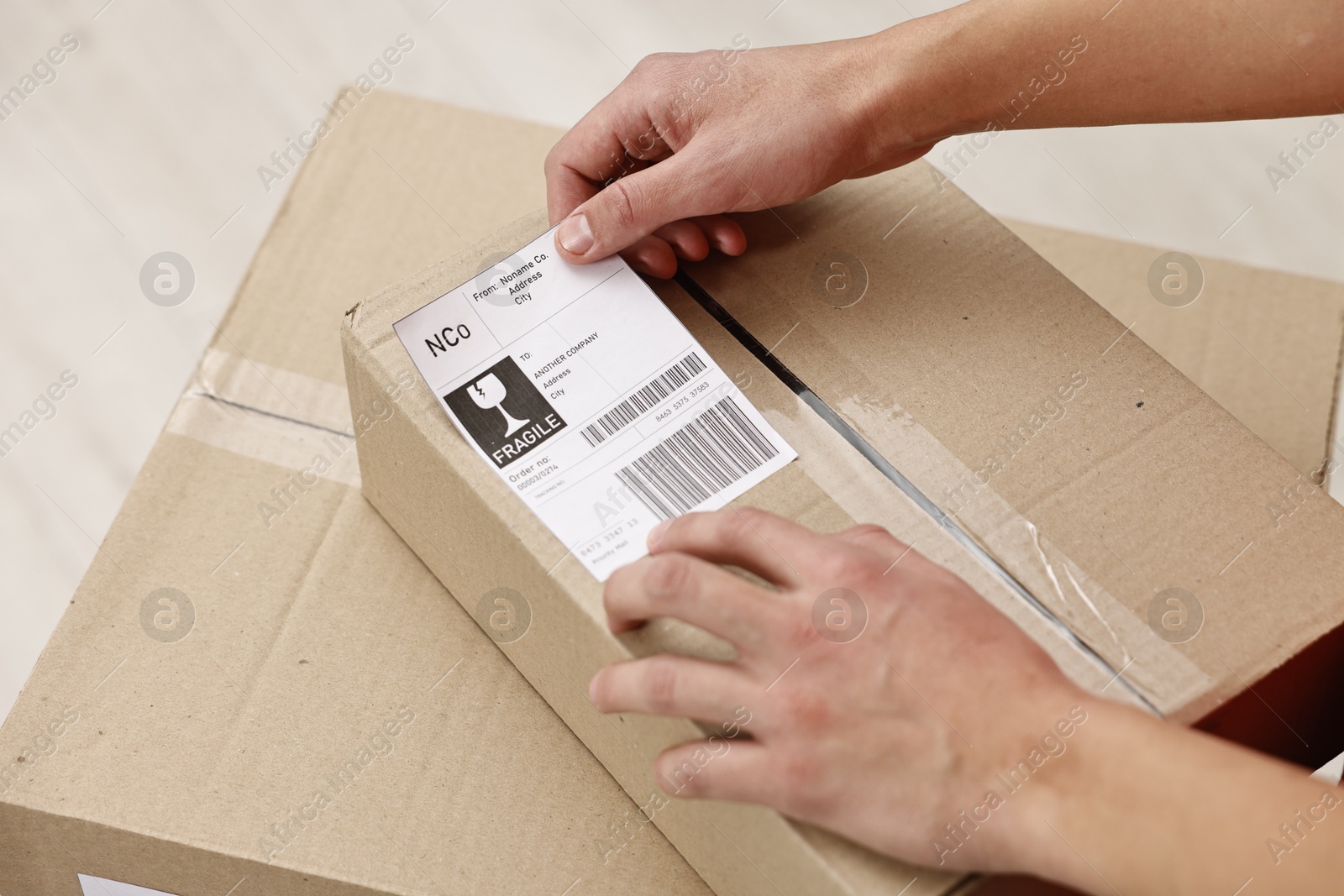 Photo of Man sticking shipping label with barcode on parcel indoors, closeup