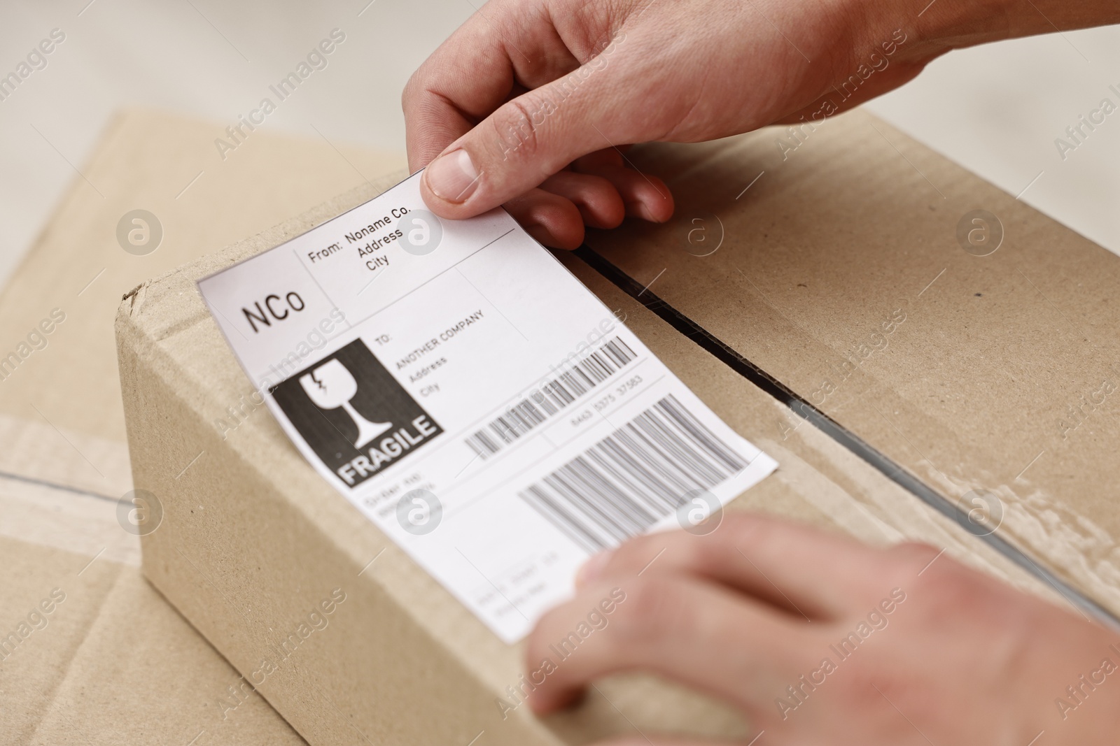 Photo of Man sticking shipping label with barcode on parcel indoors, closeup