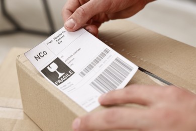 Photo of Man sticking shipping label with barcode on parcel indoors, closeup