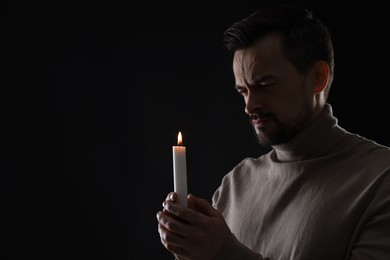 Sad man with burning candle crying on black background, space for text