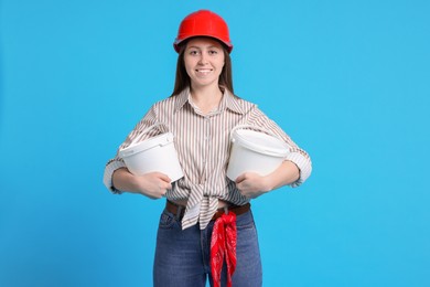 Woman wearing hardhat with buckets of paint on light blue background