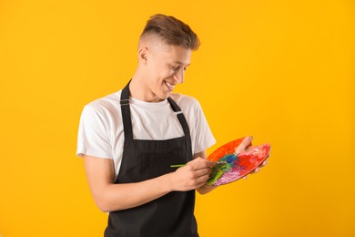 Photo of Smiling man with paintbrush mixing paints on palette against orange background