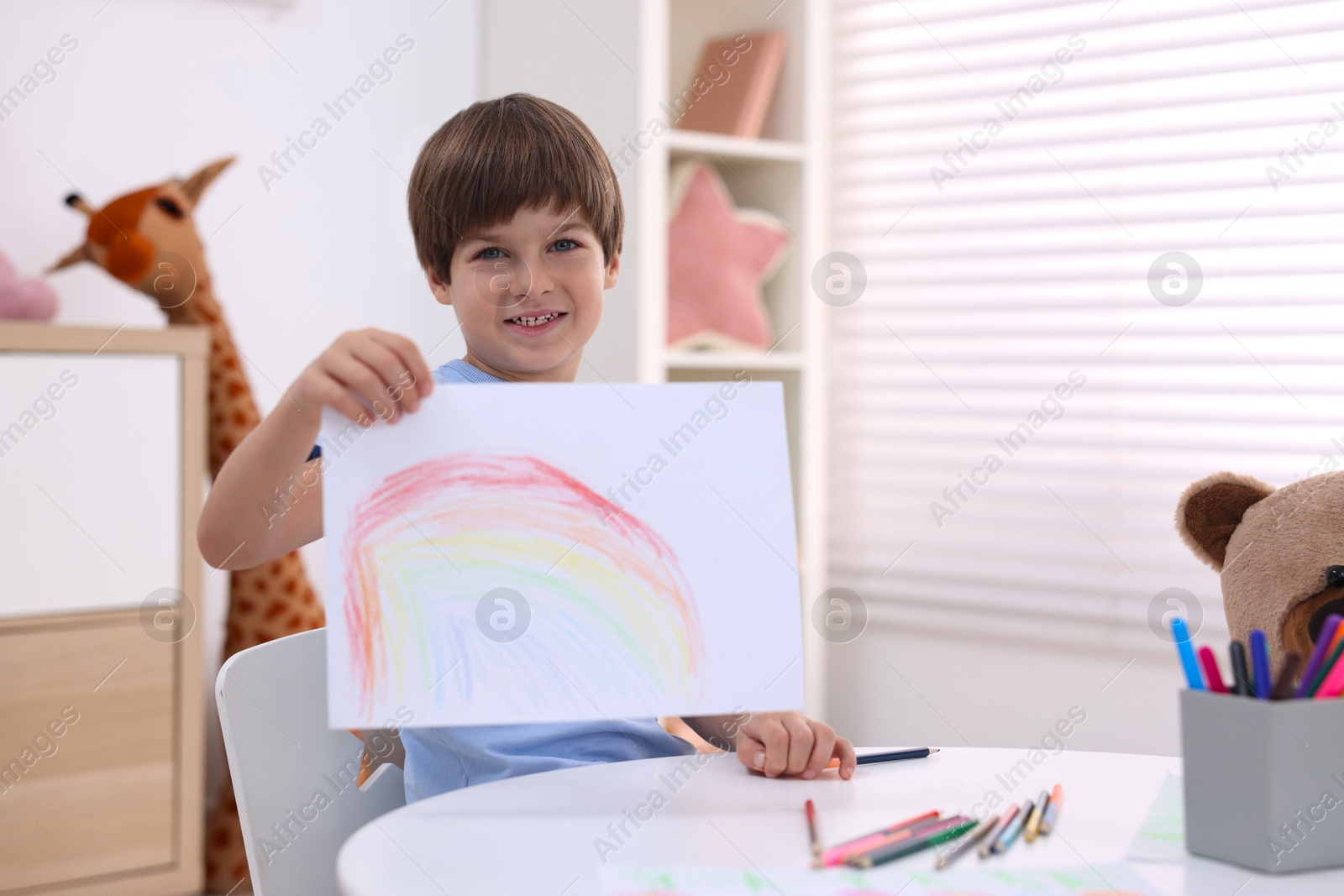 Photo of Smiling boy showing drawing at white table in kindergarten