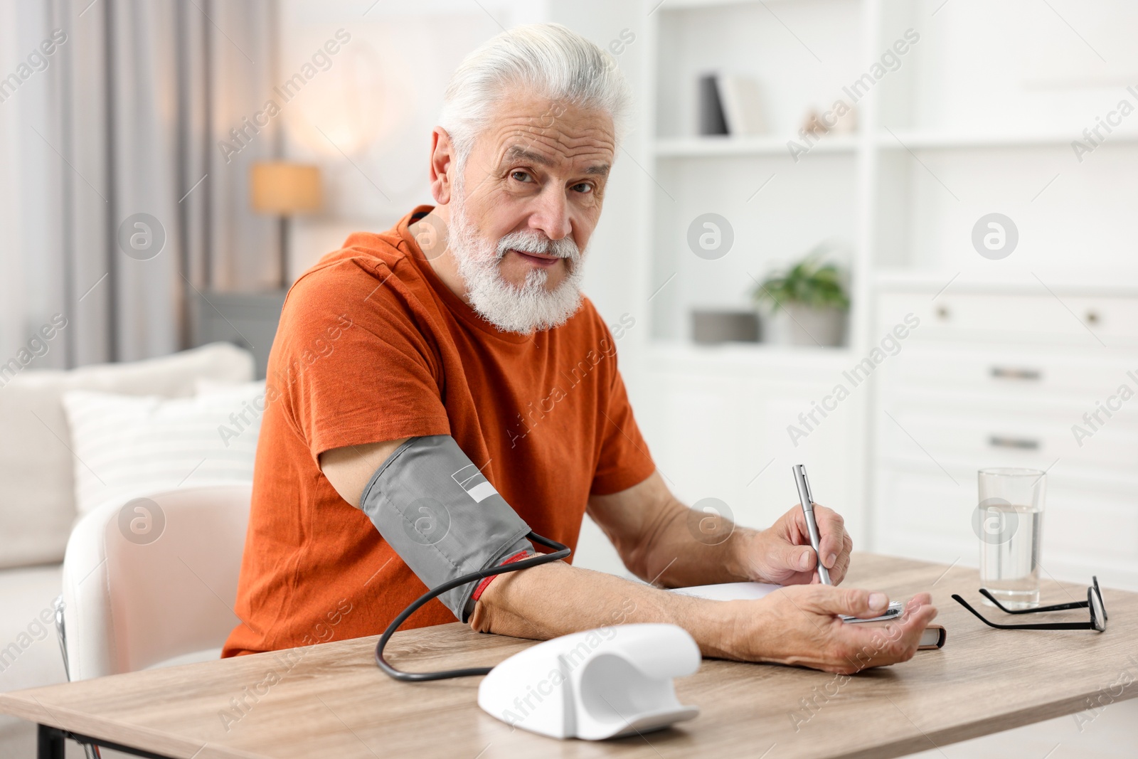 Photo of Senior man writing results of blood pressure measurement at table indoors