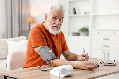 Senior man writing results of blood pressure measurement at table indoors