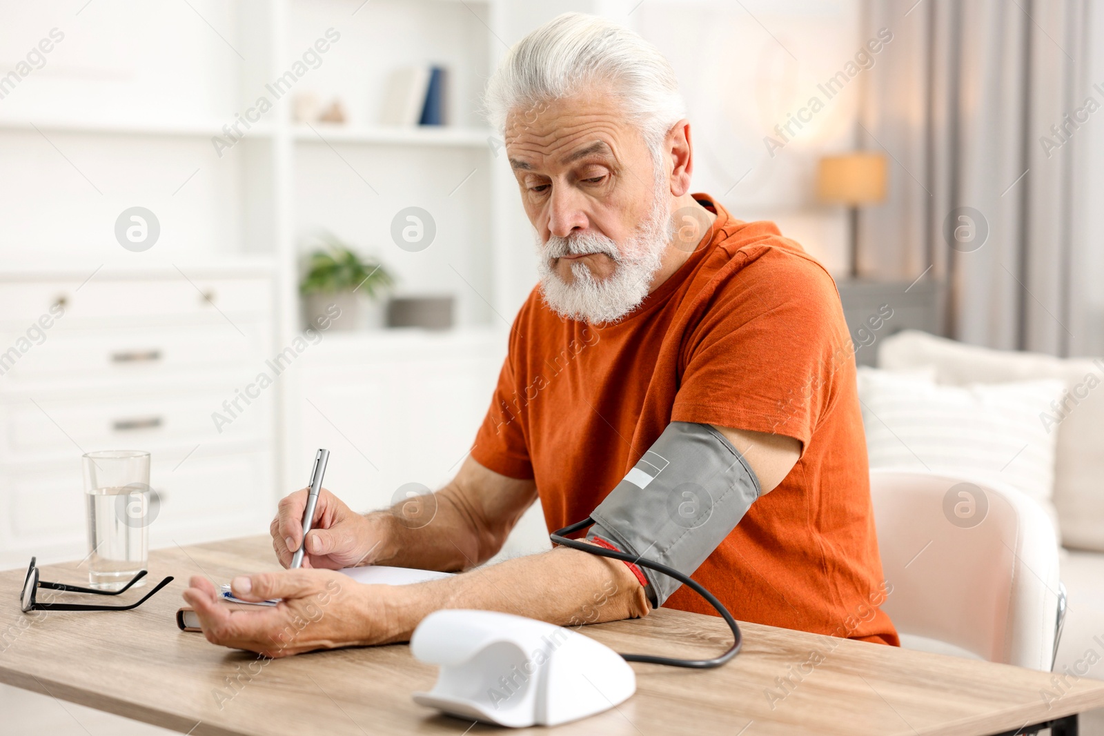 Photo of Senior man writing results of blood pressure measurement at table indoors