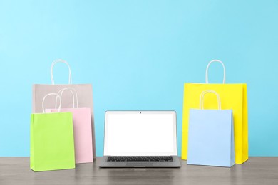 Photo of Internet shopping. Laptop and colorful paper bags on wooden table against light blue background