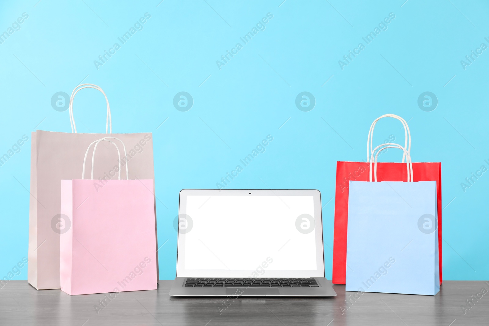 Photo of Internet shopping. Laptop and colorful paper bags on wooden table against light blue background