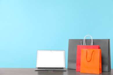 Photo of Internet shopping. Laptop and colorful paper bags on wooden table against light blue background