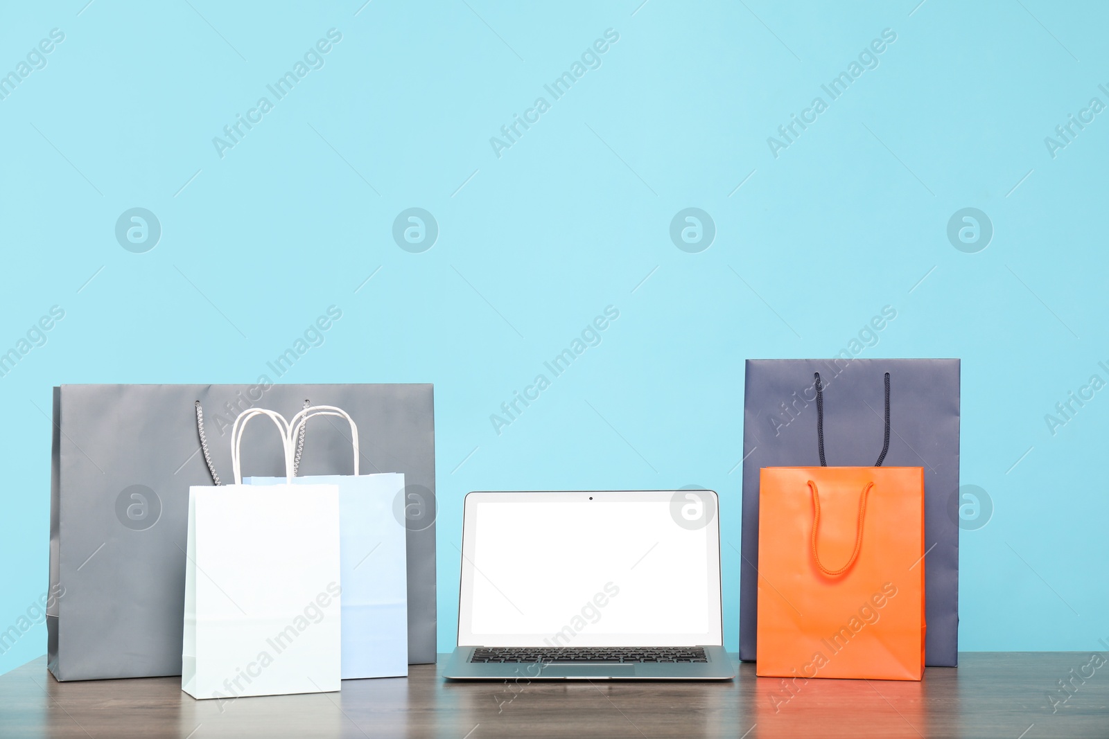 Photo of Internet shopping. Laptop and colorful paper bags on wooden table against light blue background