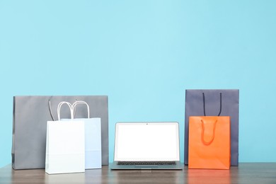 Photo of Internet shopping. Laptop and colorful paper bags on wooden table against light blue background