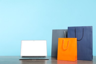 Photo of Internet shopping. Laptop and colorful paper bags on wooden table against light blue background