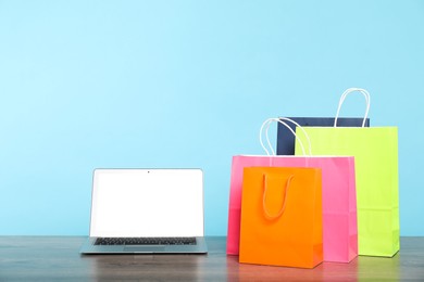 Photo of Internet shopping. Laptop and colorful paper bags on wooden table against light blue background