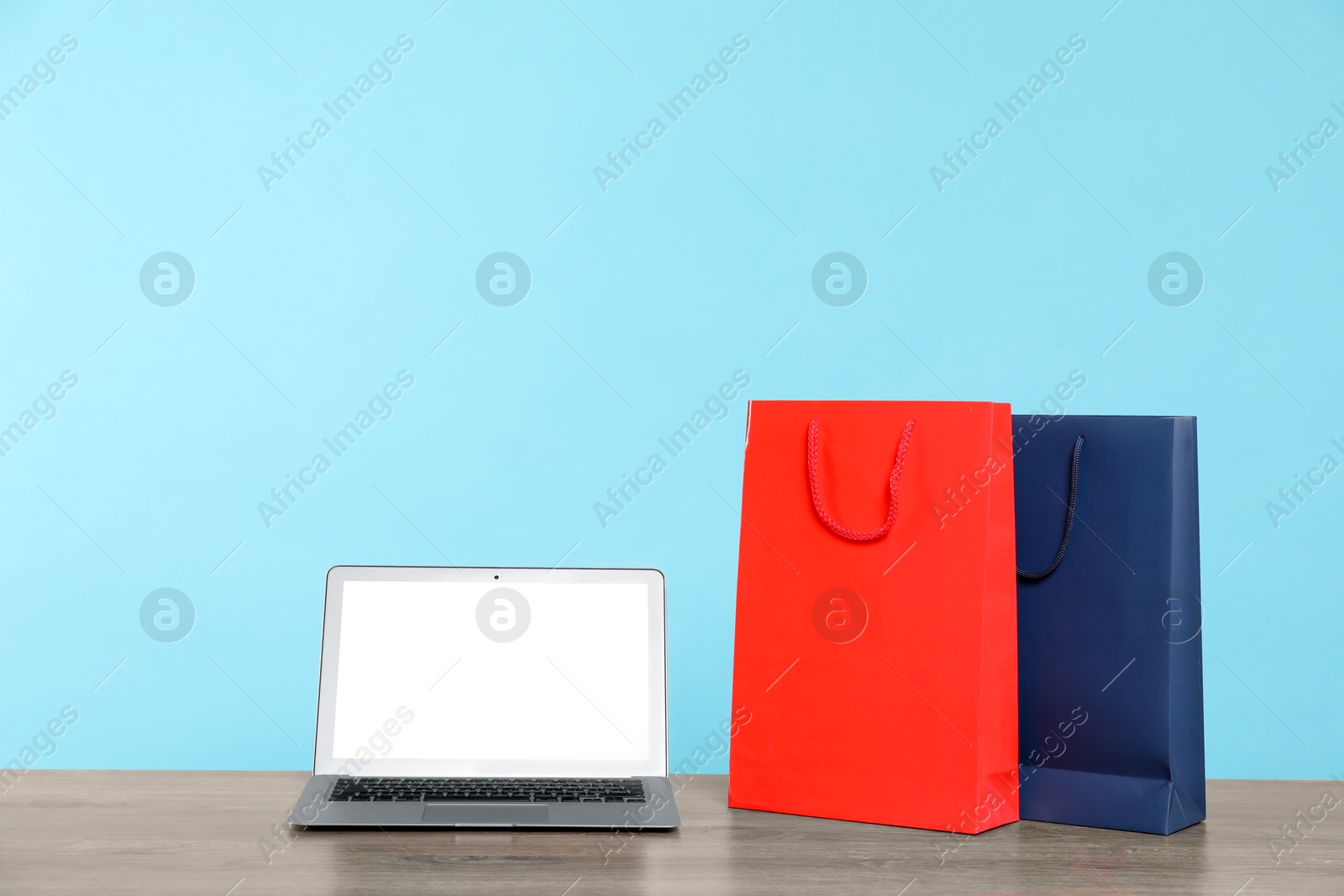 Photo of Internet shopping. Laptop and colorful paper bags on wooden table against light blue background