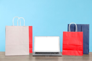 Photo of Internet shopping. Laptop and colorful paper bags on wooden table against light blue background