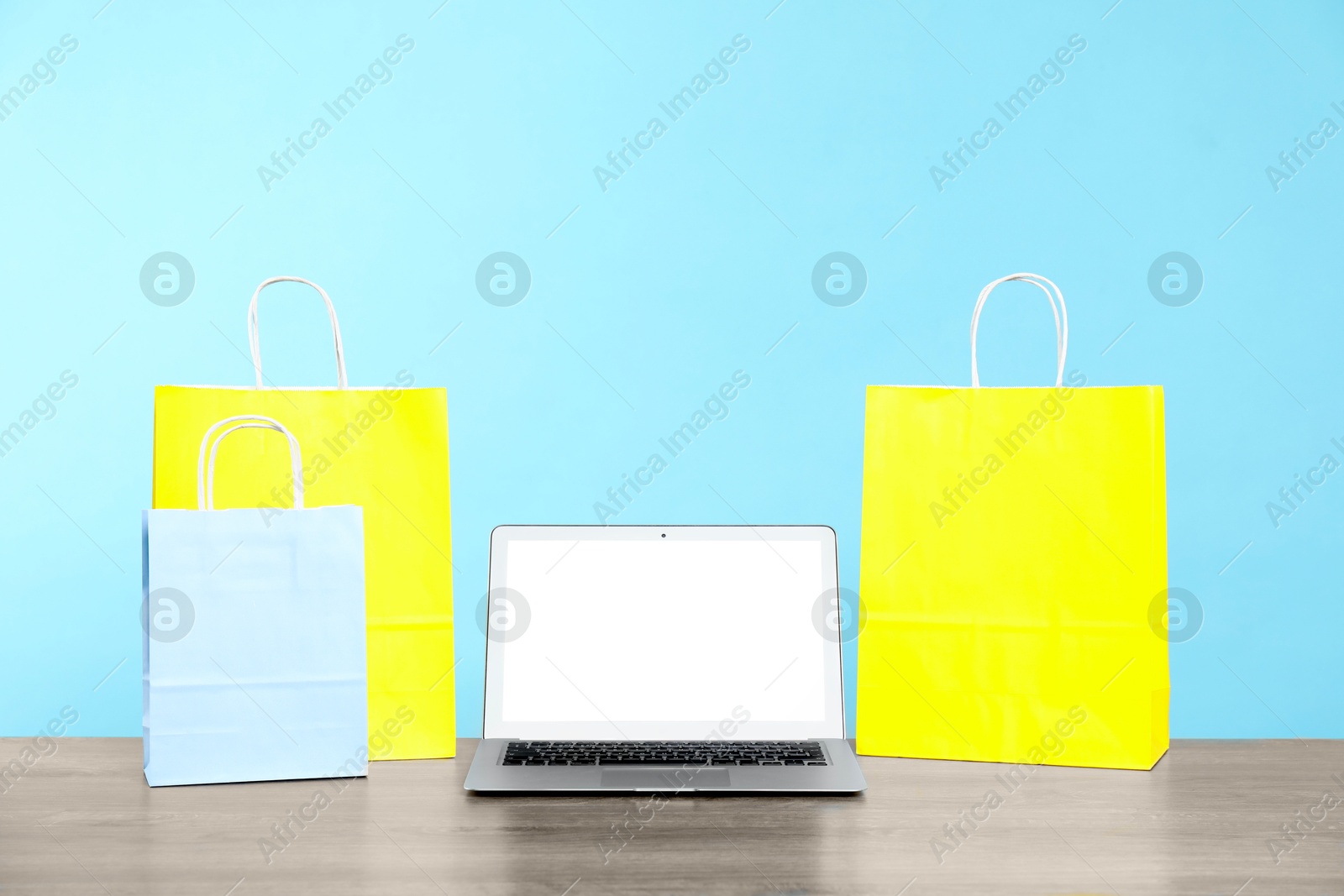 Photo of Internet shopping. Laptop and colorful paper bags on wooden table against light blue background