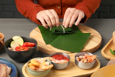 Photo of Woman tying banana leaf with food at wooden table with products, closeup. Healthy eco serving