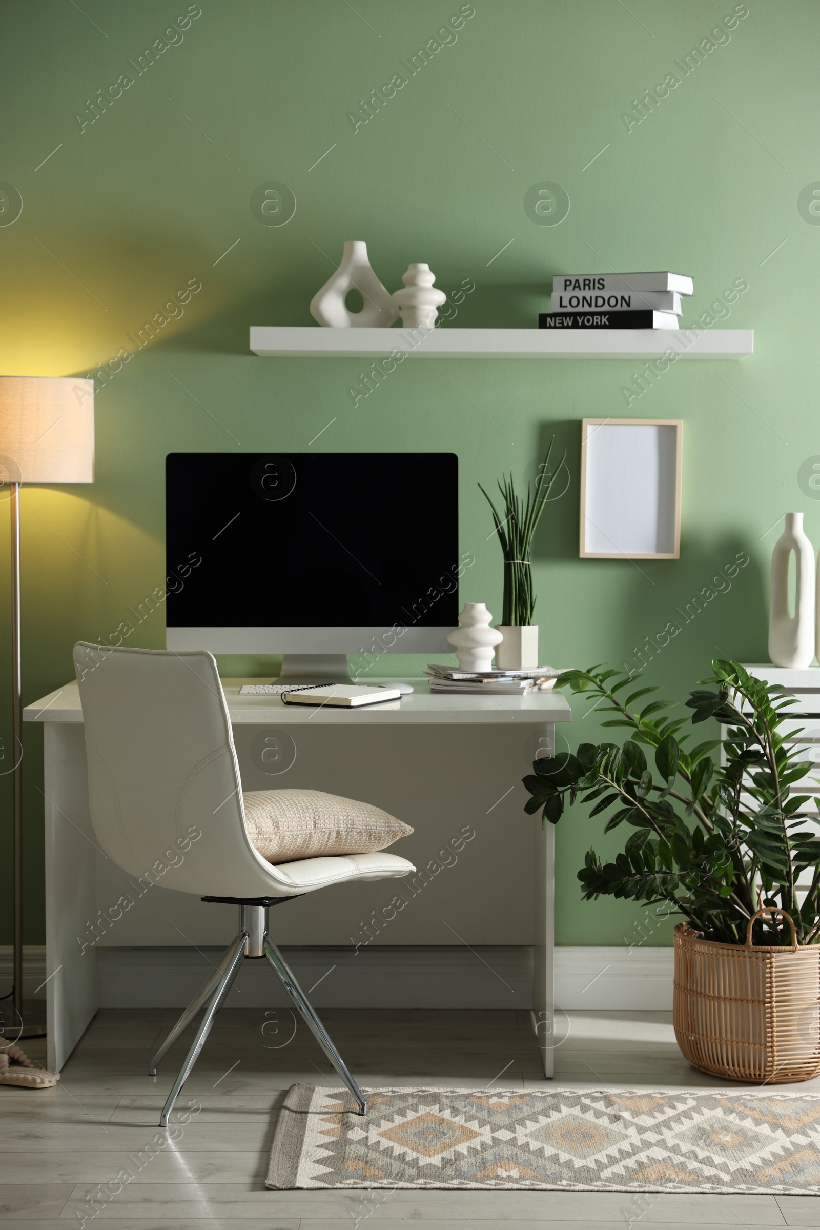 Photo of Home office. Comfortable workplace with desk, computer and lamp near light green wall in room