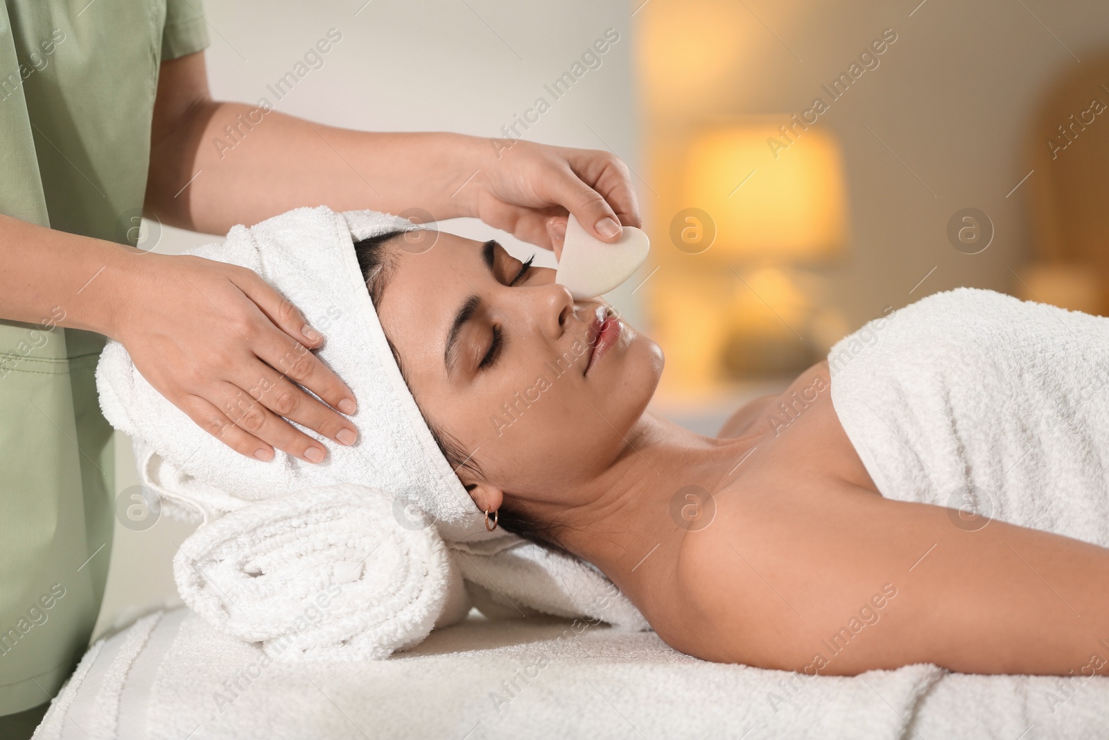 Photo of Young woman receiving facial massage with gua sha tool in spa salon