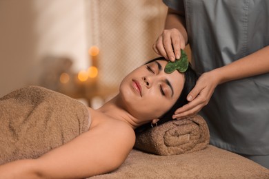 Photo of Young woman receiving facial massage with gua sha tool in spa salon