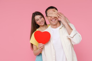 Photo of Lovely woman surprising her boyfriend with paper heart on pink background, space for text. Valentine's day celebration