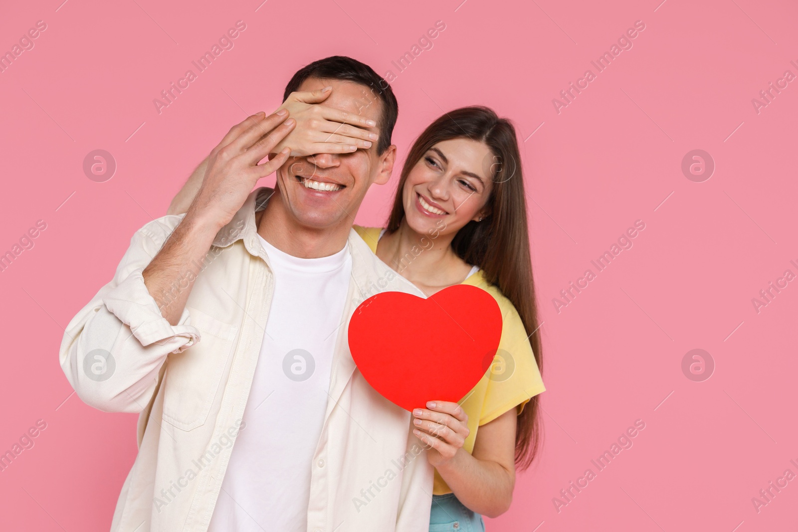 Photo of Lovely woman surprising her boyfriend with paper heart on pink background, space for text. Valentine's day celebration