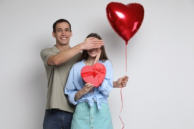 Lovely man surprising his girlfriend with Valentine's day gift and heart shaped balloon on light background