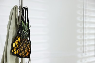 Photo of Macrame bag with fruits and jacket on clothing rack near white wall. Space for text