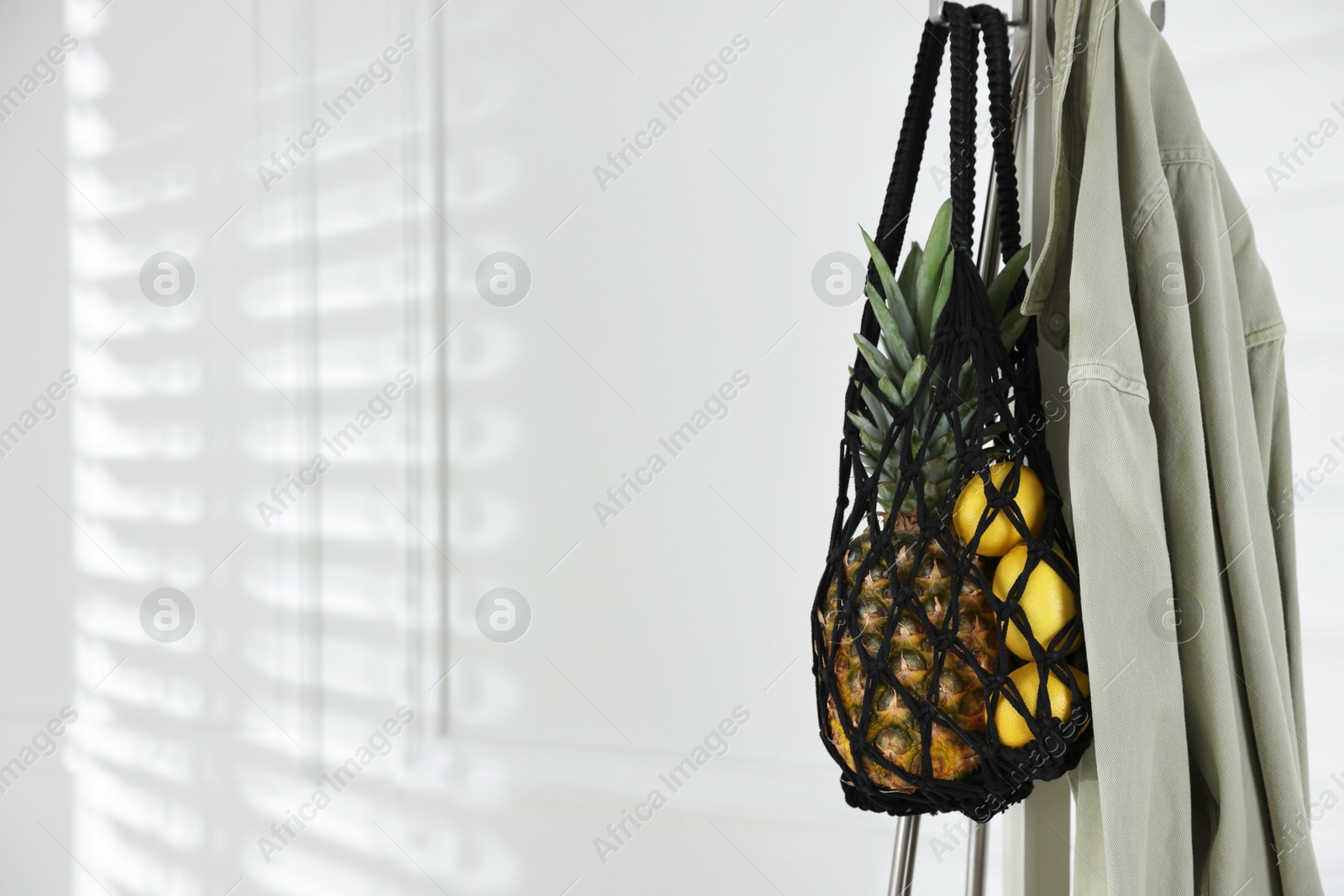 Photo of Macrame bag with fruits and jacket on clothing rack near white wall. Space for text