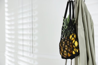 Photo of Macrame bag with fruits and jacket on clothing rack near white wall. Space for text