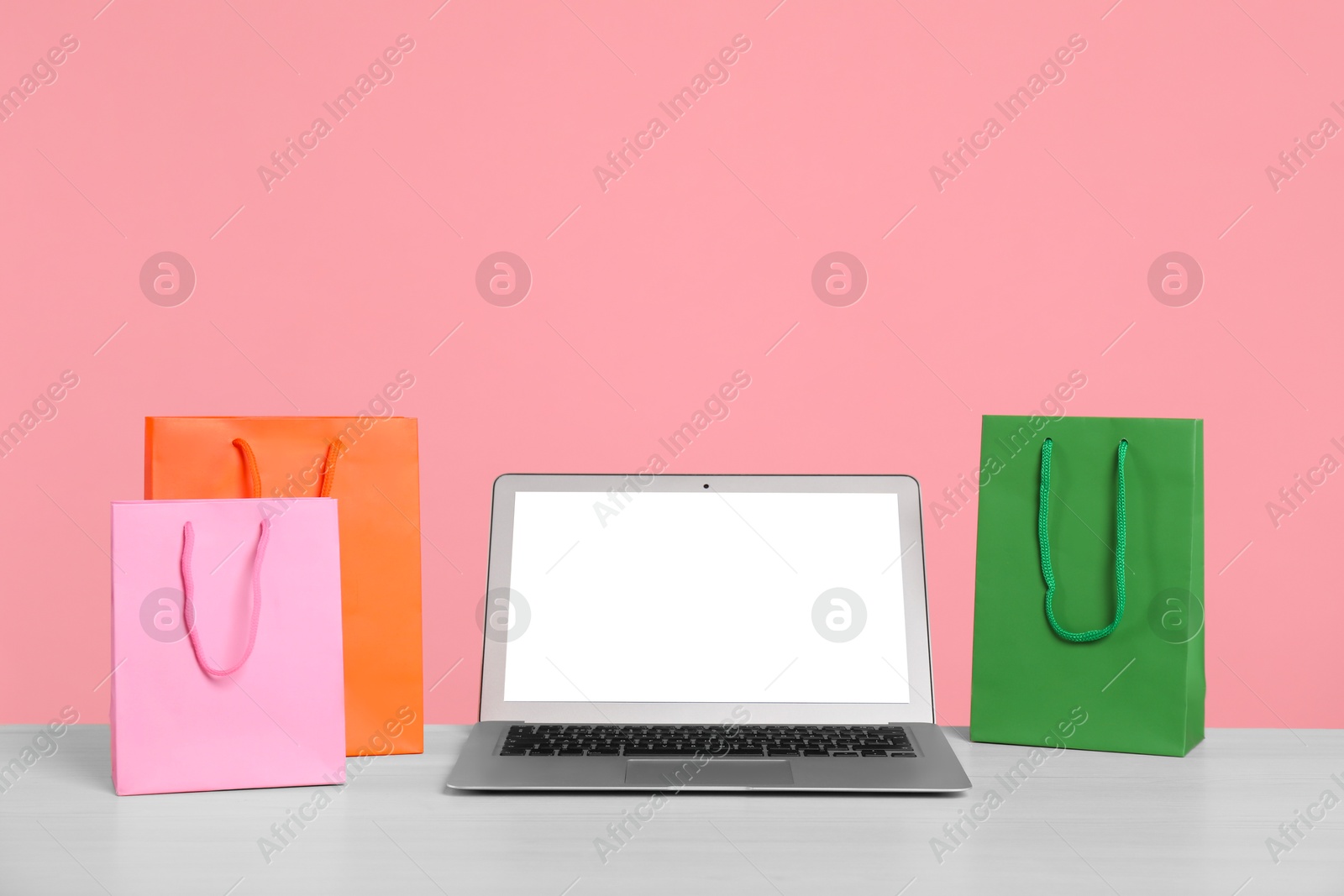 Photo of Internet shopping. Laptop and colorful paper bags on table against pink background