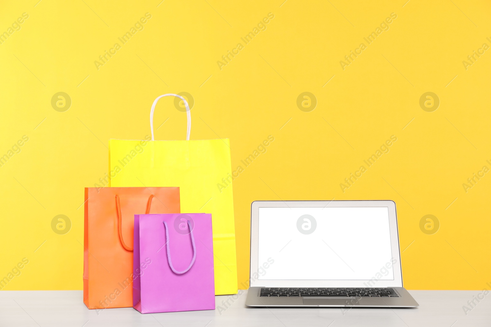 Photo of Internet shopping. Laptop and colorful paper bags on table against yellow background