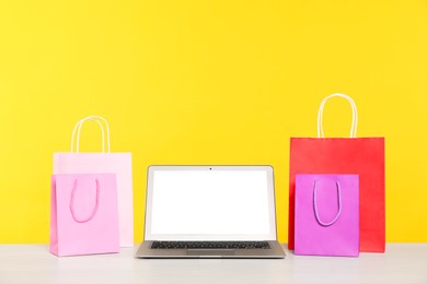 Photo of Internet shopping. Laptop and colorful paper bags on table against yellow background