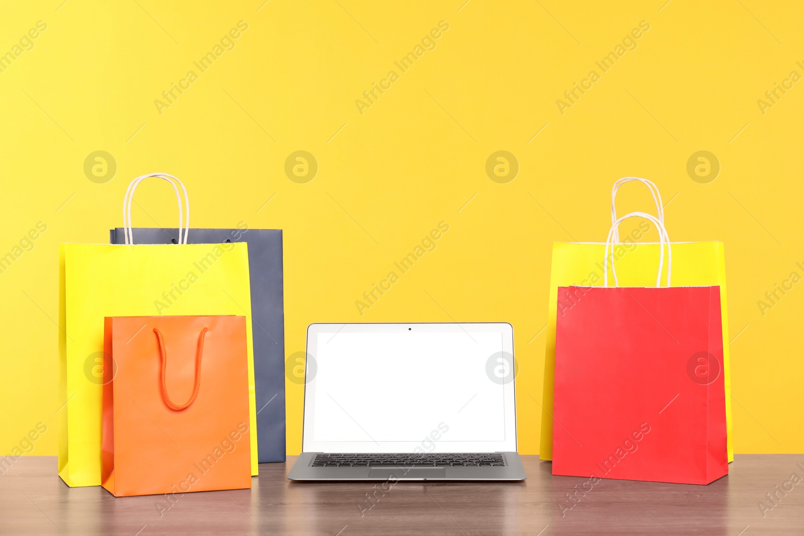 Photo of Internet shopping. Laptop and colorful paper bags on wooden table against yellow background