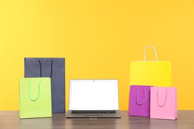 Photo of Internet shopping. Laptop and colorful paper bags on wooden table against yellow background