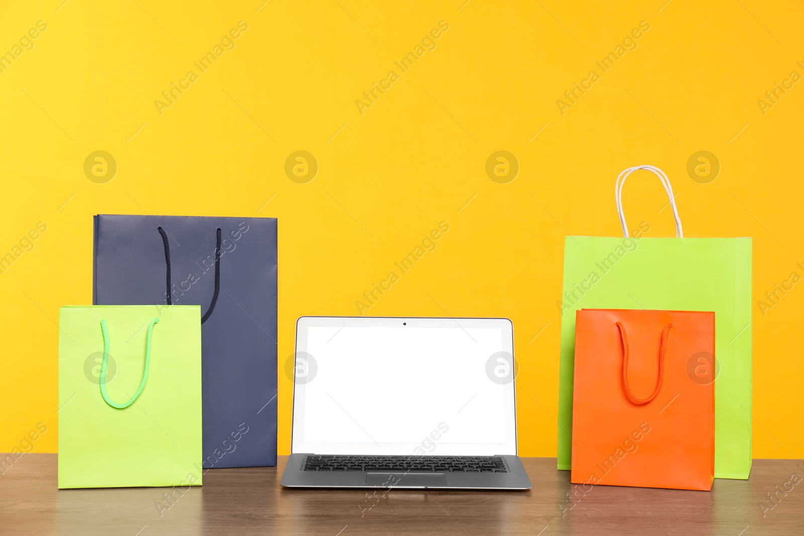 Photo of Internet shopping. Laptop and colorful paper bags on wooden table against yellow background